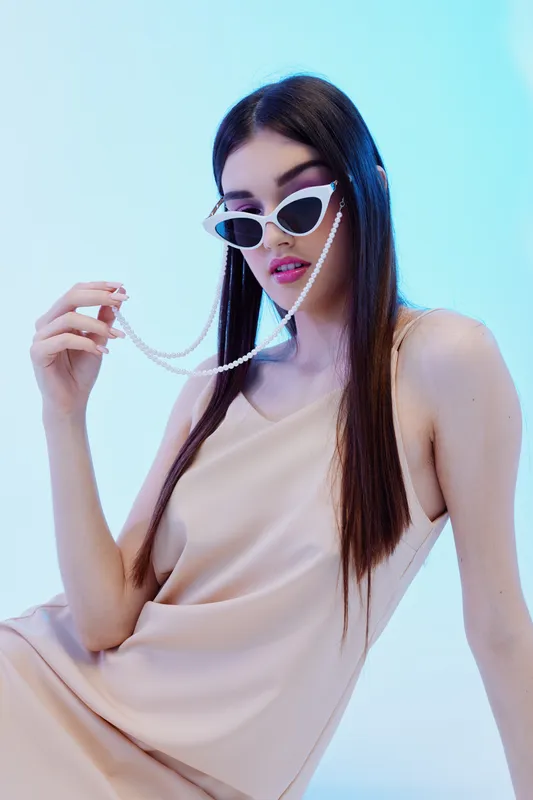 Young woman in a beige dress wearing white sunglasses with a pearl chain poses against a blue background. Concept of beauty, fashion, elegance, and modern style.