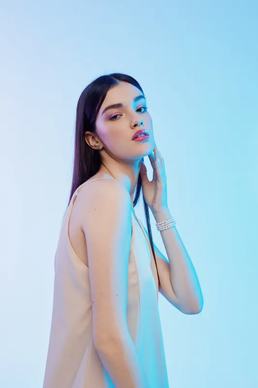 Young woman with long dark hair poses in a sleeveless top against a blue background. Soft lighting highlights her beauty and fashion style in a modern studio setting.