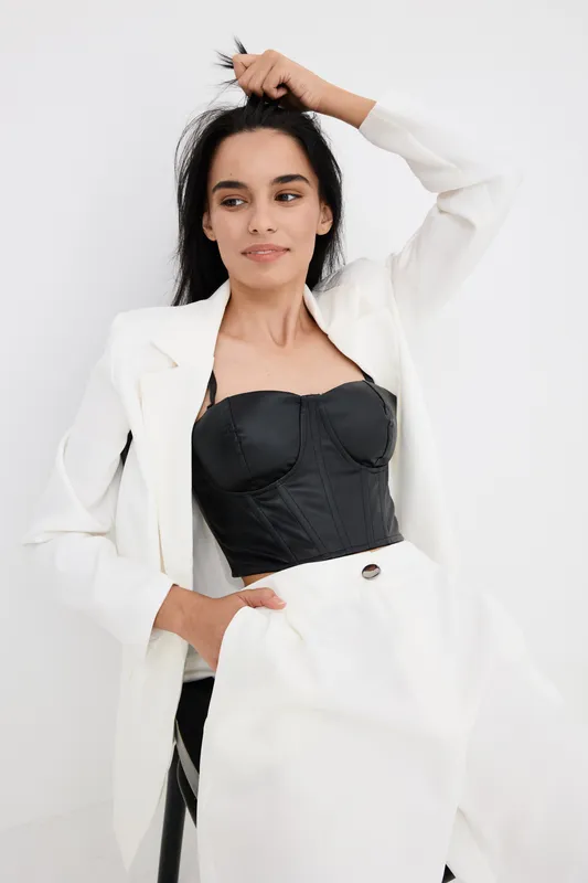 Young woman with black corset and white blazer posing confidently indoors, showcasing modern fashion style and graceful elegance in minimalistic bright setting.
