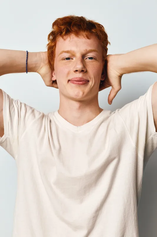 Young man with red hair in casual white t shirt stands relaxed with hands behind head against neutral background, confident and calm expression on face.