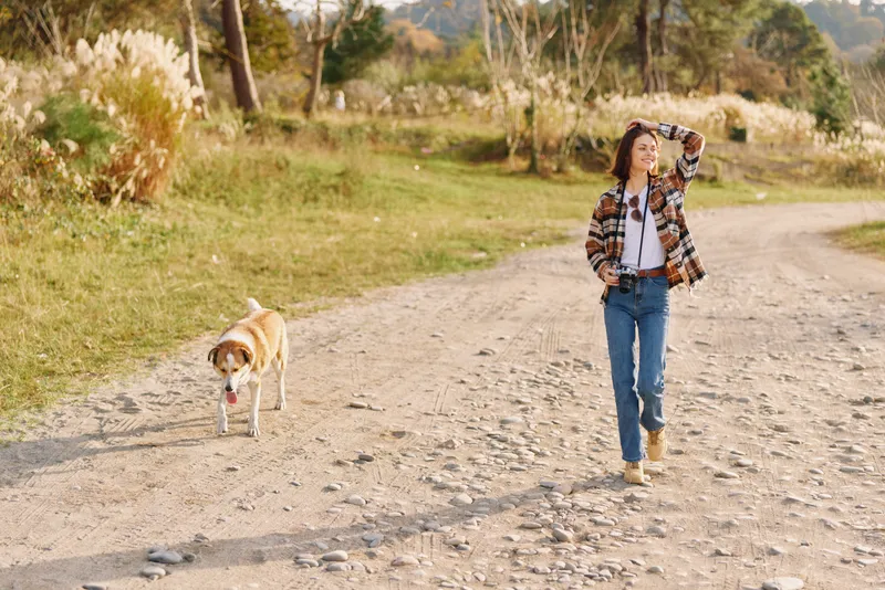 Woman walking with her dog along a rocky dirt path in a park during autumn, casual outfit, enjoying nature and fresh air on a sunny day, capturing outdoor activity and a peaceful moment.