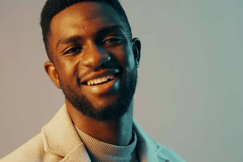 Smiling African man in a light sweater and jacket against a soft blue background, showcasing positive emotions and warmth