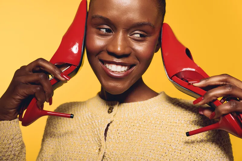 Smiling dark skinned African woman holds red high heels near her ears on yellow background. Concept of fashion, beauty, joyful emotions, and stylish accessories for women.