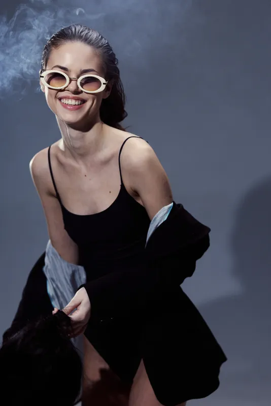Smiling woman wearing retro sunglasses and black dress, enjoying casual moment with confident expression.