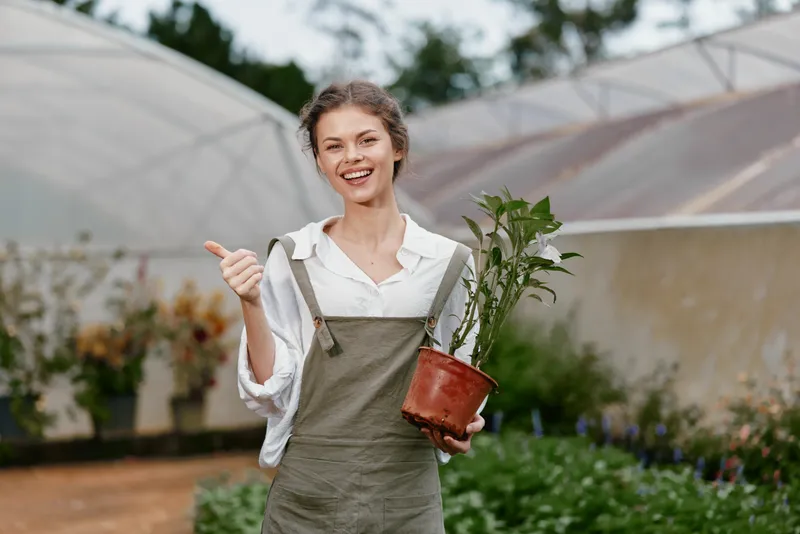 Happy young woman in green apron holding a potted plant, expressing joy and positivity in a greenhouse, symbolizing gardening and nature
