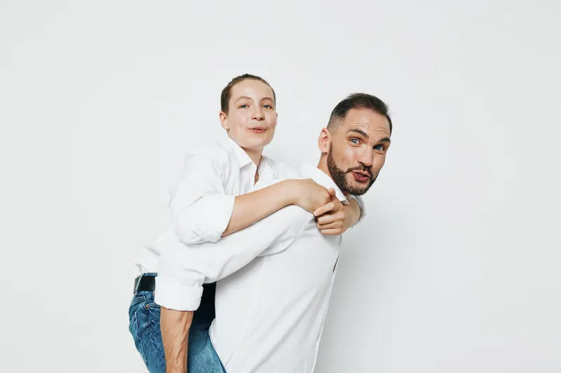 A professional man with a casual smart look carries a colleague on his back against a clean white background, portraying teamwork, confidence, and positive energy.