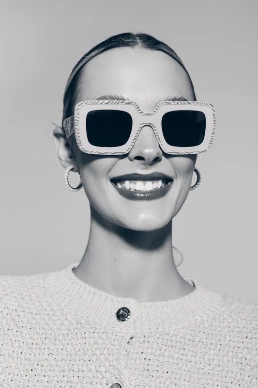 Smiling white woman with pale skin wearing large sunglasses and hoop earrings in studio. Concept of fashion accessories and confident style.