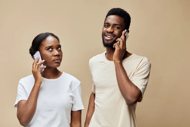 Young African couple communicating on smartphones, showcasing a mix of curiosity and joy, dressed in casual clothing against a soft beige background