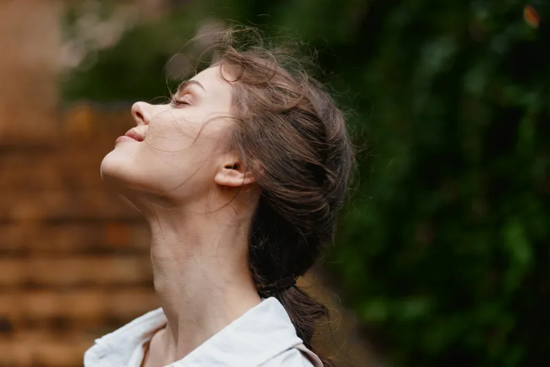 Young woman with brown hair in a white shirt enjoying the outdoors, exuding joy and tranquility against a lush green background in a springtime setting