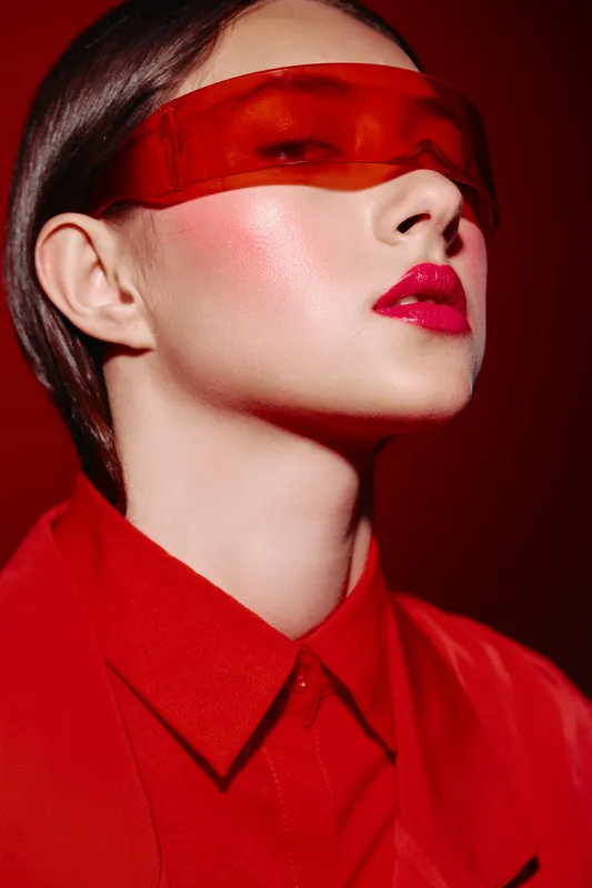 Stylish woman with glossy red lipstick and shimmer cheek makeup wears translucent red visor. Portrait on dark red background captures bold beauty, creative fashion concept, modern style elegance.
