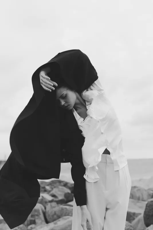 Monochrome scene of two figures on a rocky shore, one in a dark veil embracing the other, capturing contrast, mood, and quiet mystery.