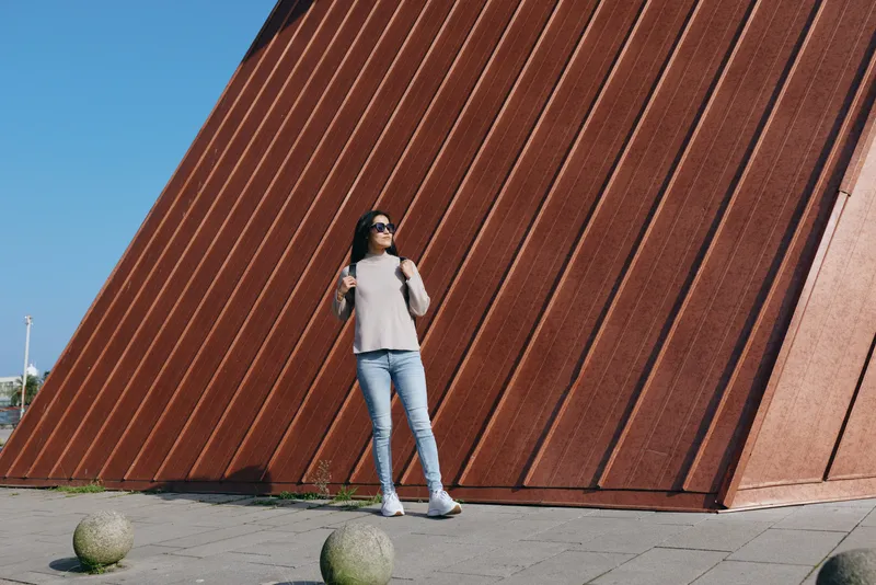 Woman stands in casual attire beside a tall rust colored wall, outdoor urban setting with blue sky, relaxed pose, contemporary architecture, street style and modern lifestyle
