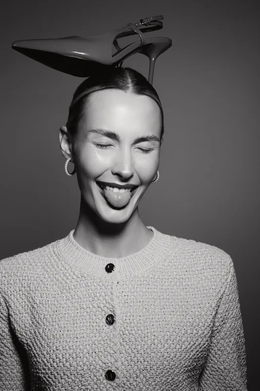 Smiling white woman with light skin and short hair sticks out tongue, wearing textured sweater and high heel shoe on head in studio portrait.