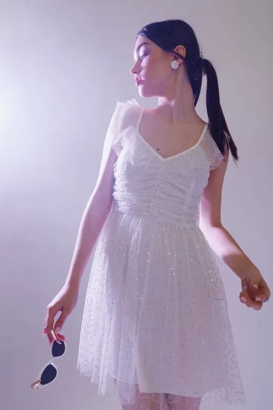 Young woman in a white dress holding sunglasses looks to the side. Soft lighting highlights her beauty and fashion style. Concept for elegance, youth, and summer wear.