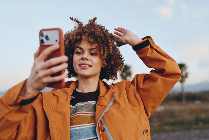 Woman with curly hair takes a selfie with a phone outdoors, smiling in an orange jacket. Close view lifestyle moment in fresh daylight, relaxed pose against a soft sky.