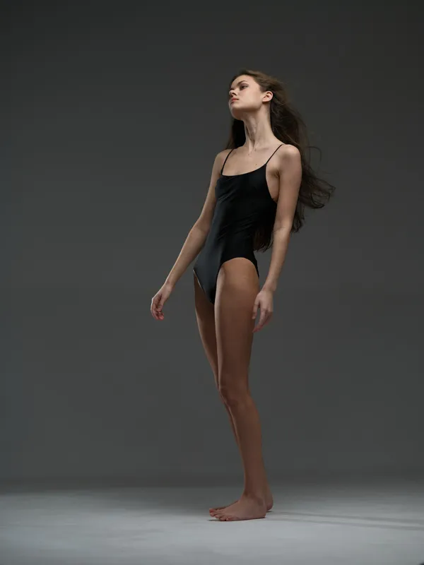 Slim woman in black bodysuit standing on gray background with confident pose and calm expression, elegant minimalism style
