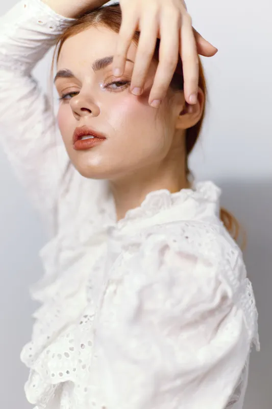 Elegant young woman in a white blouse expressing confidence and beauty against a soft neutral background, showcasing a modern and minimalist fashion style