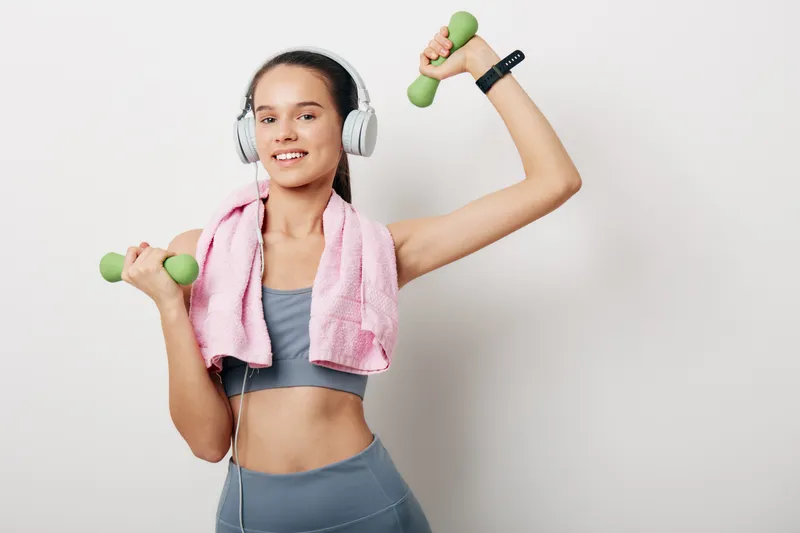 Young fit woman in gray sportswear with a pink towel around her neck lifts green dumbbells while wearing headphones and a smartwatch against a clean white background, smiling.