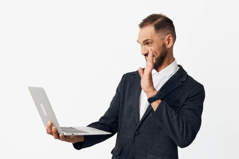 A professional man stands on a solid colored background, holding a laptop and showing thoughtful, confident emotion as he engages with the screen.