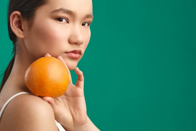 Asian woman holds an orange near her shoulder against a vivid green background, creating a fresh, simple portrait. The image conveys natural beauty, health, and calm confidence with a bright,