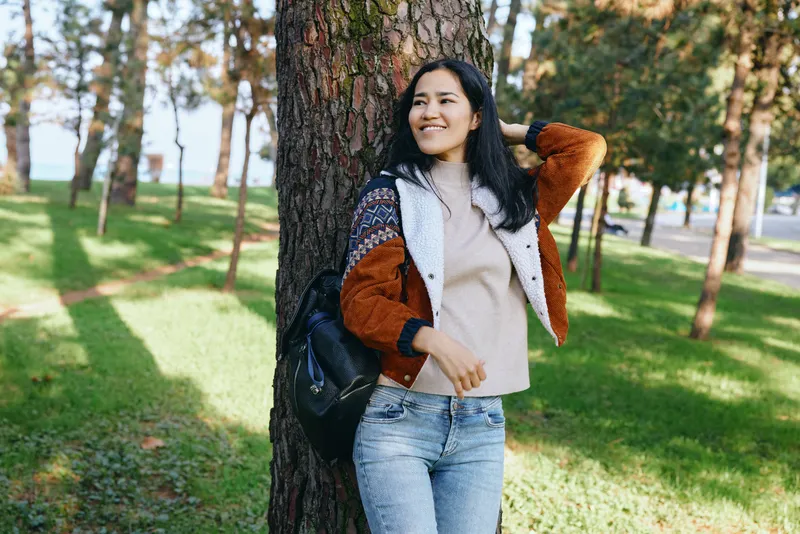 Smiling woman leans against a tree in a sunlit park, wearing a color block jacket and jeans, enjoying a relaxed outdoor moment amid green grass and warm light.