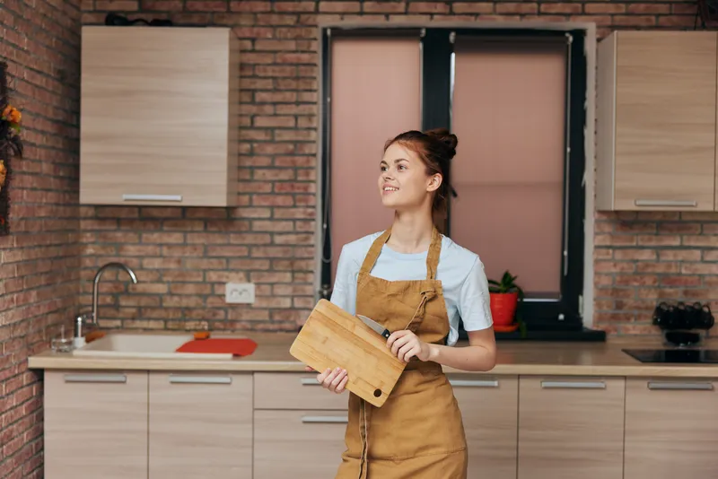 Красивая женщина в фартуке стоит с разделочной доской и ножом на кухне