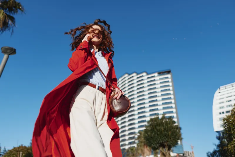 Confident woman walking outdoors in red coat and white pants, sunny day with clear blue sky, modern urban background, casual style, joyful expression