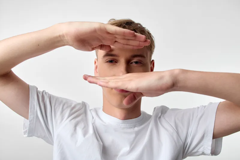 Frame hands create a geometric boundary around a man in a white tee, neutral background, capturing candid creative framing and simple studio style.