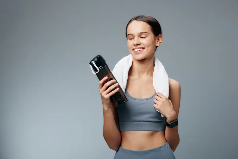 Fit young woman in workout clothes holds a water bottle and wears a towel over her shoulders, smiling after exercise in a minimalist studio with soft gray background lighting.