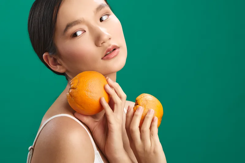 asian woman holds oranges against a vivid green backdrop, soft lighting enhances skin tones, cheerful expression, casual pose suggesting freshness and vitality, creating a bright and inviting mood