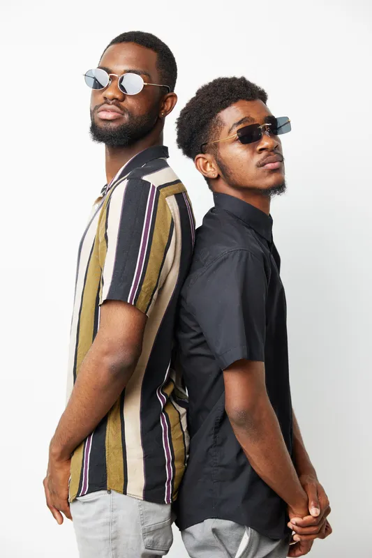 Two stylish black men stand back to back, showcasing modern fashion with sunglasses. Their confident poses and distinct outfits highlight urban culture and camaraderie.