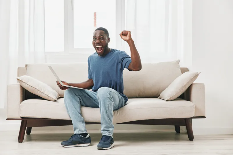 Excited young man celebrating success while sitting on a couch with a laptop, conveying joy and enthusiasm in a bright, modern living room