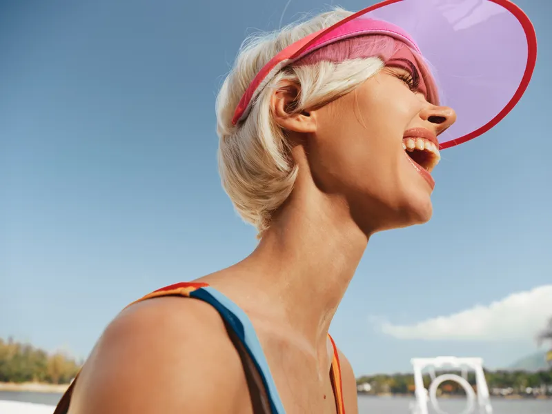 Bright portrait of a joyful woman wearing a pink visor, laughing under a clear blue sky. Sunlit skin and casual summer attire capture carefree optimism.
