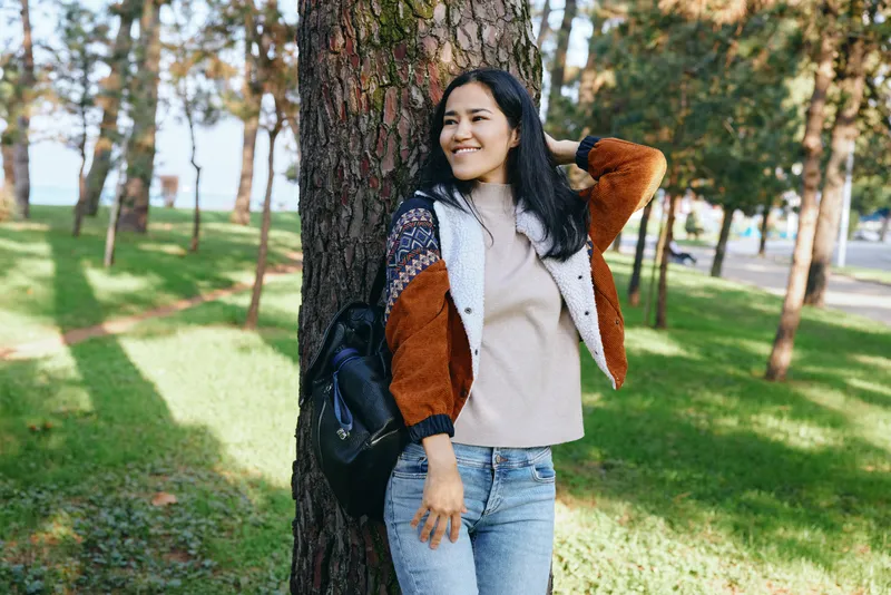 Woman leaning against a tree in a sunny park, wearing a cozy sweater and denim jeans, with a backpack, enjoying outdoors and relaxed style in natural scenery