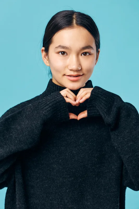 Woman posing with hands forming heart shape near chest, wearing black sweater on blue background. Portrait of young Asian female with calm expression and casual outfit in studio.