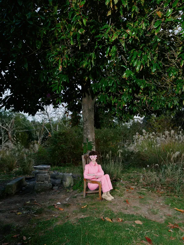 A woman sitting under tree in a garden wearing a pink dress enjoying a tranquil outdoor moment in natural shade, with calm atmosphere, soft light and casual seating.