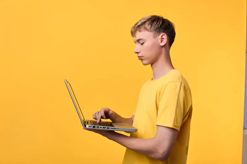 Laptop held by a yellow shirted man in profile against a bright yellow backdrop, focused on digital work, technology use, and learning in a modern studio setting.