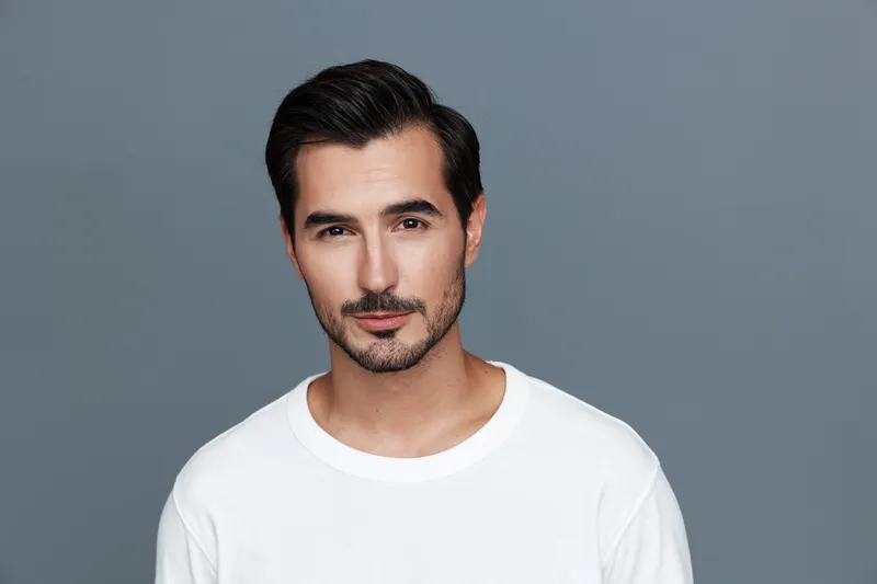 Stylish man with short dark hair wearing a white t-shirt looks confidently at the camera against a gray background, exuding charisma and charm.