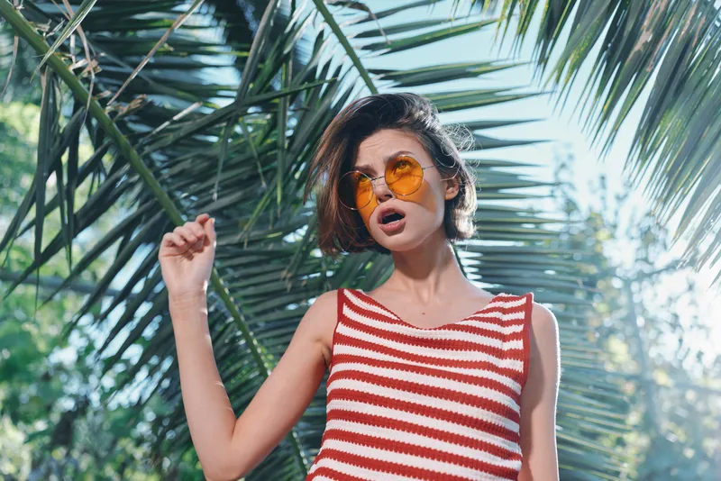 A stylish woman wearing a striped red and white top stands outdoors among palm leaves, posing confidently in sunny weather, radiating vibrant fashion and carefree energy.