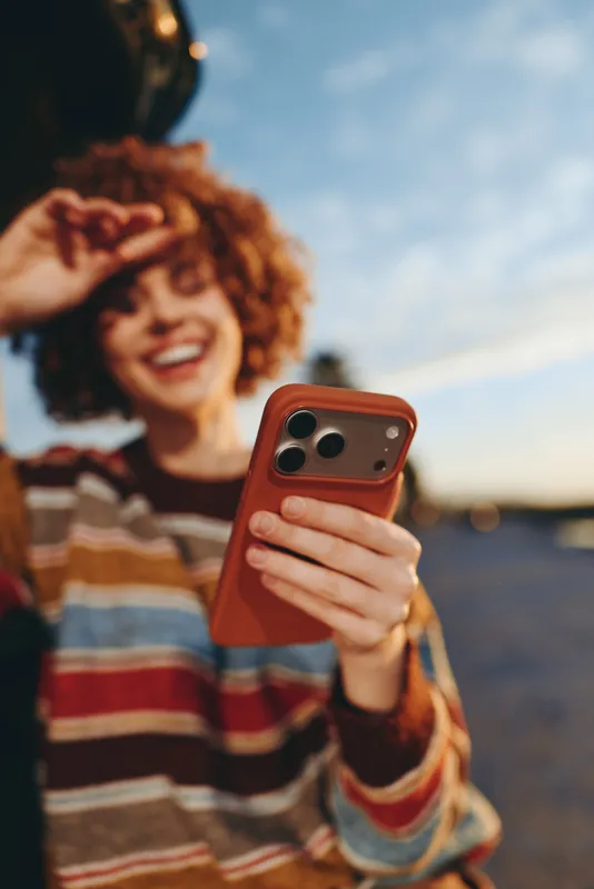 Woman lifestyle portrait outdoors, curly hair, striped sweater, holding smartphone for a selfie, smiling toward camera against sunset sky, shallow depth of field, street bokeh light