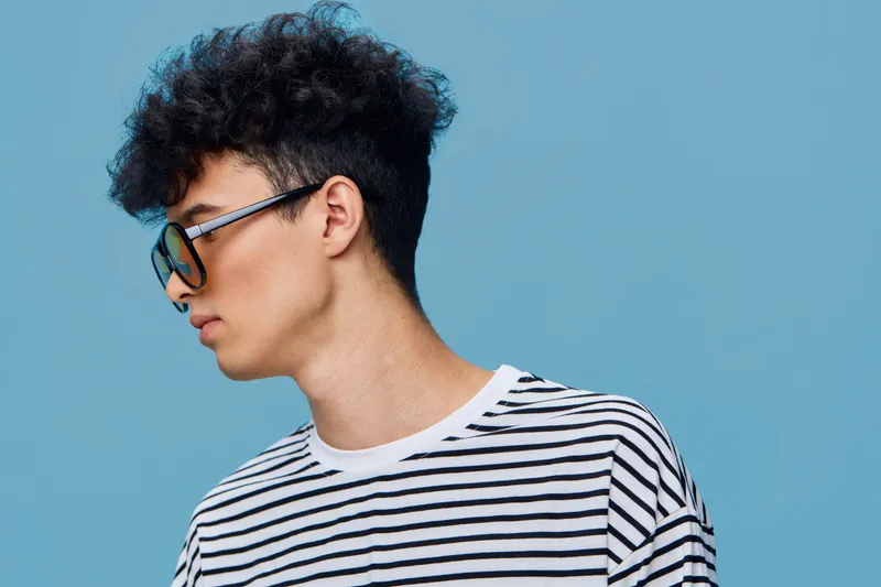 young man with curly hair wearing sunglasses and striped shirt on blue background, casual style and modern look with side profile view