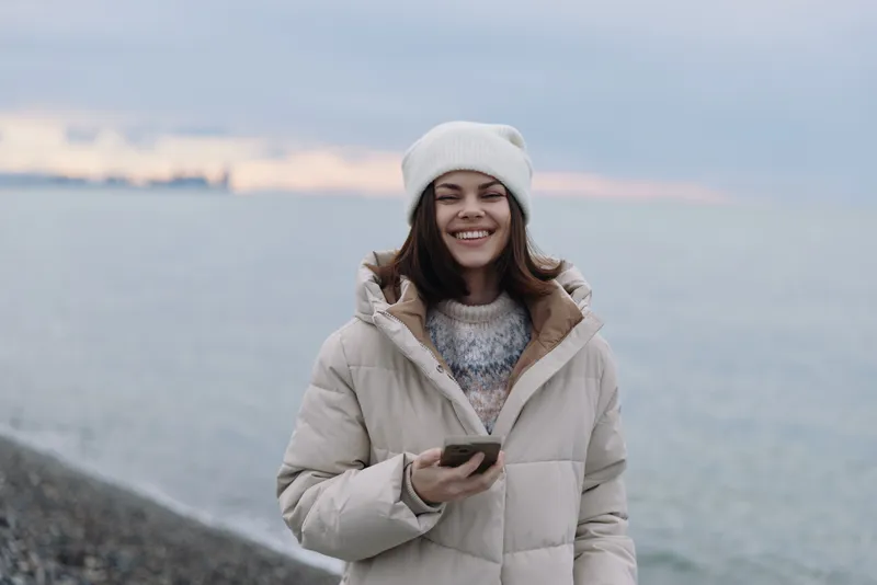 Winter portrait of a smiling woman in a padded coat and knit hat, standing by a calm coastline with a phone in hand, soft sunset light and rocky shore.