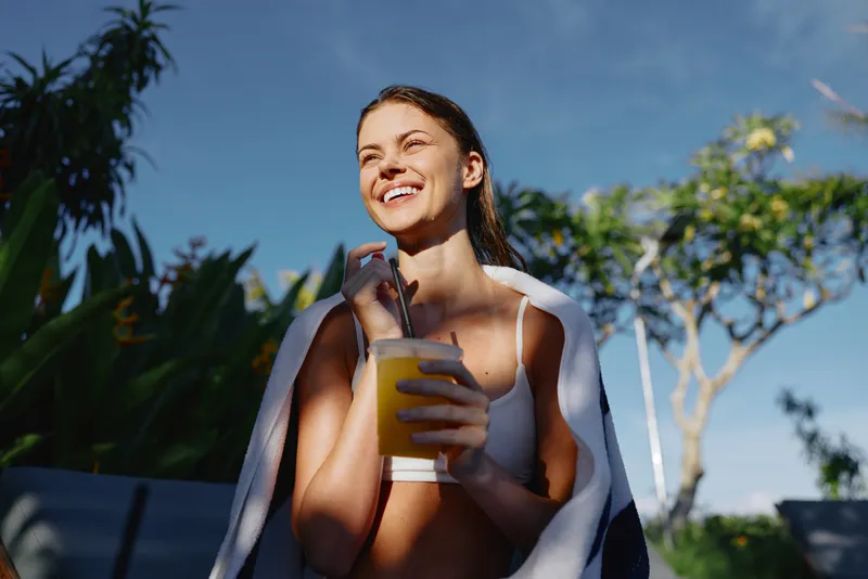 Happy woman outdoors enjoys a cold drink under sunny skies, wearing casual swimwear with a towel draped over her shoulders, capturing a relaxed lifestyle moment in a tropical setting, bright mood and