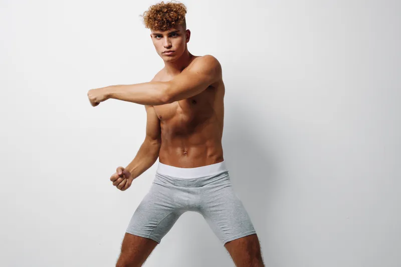 Young man with curly hair posing shirtless in gray shorts against white background, showing athletic build and confident expression in studio lighting for fitness or fashion concept.