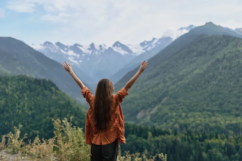 Person stands with arms raised at a scenic mountain viewpoint, enjoying a vast forested landscape and snowcapped peaks in the distance, radiating freedom and awe