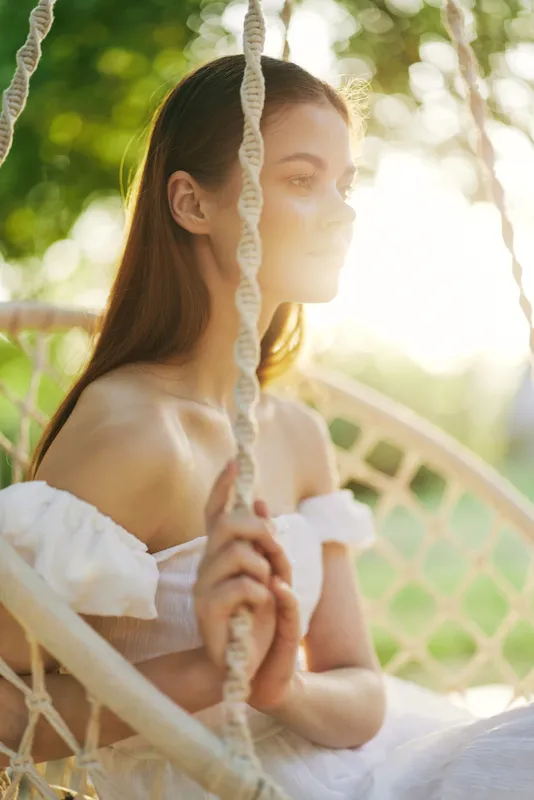 A serene young woman in a white dress on a swing, basking in warm sunlight, evoking tranquility and natural beauty in a vibrant outdoor setting