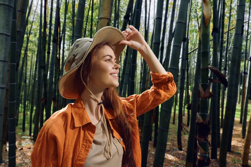 Woman in hat smiling in a bamboo forest, portrait of a happy redhead outdoors exploring nature with sunlight through stalks, casual jacket and straw hat for travel and adventure.