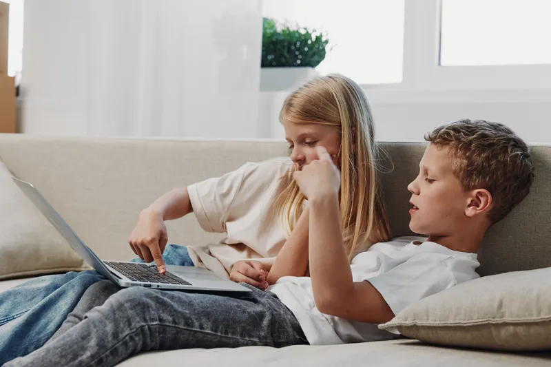 Two children sitting on a sofa using a laptop together in a bright living room. The boy and girl focus on the screen, sharing a moment of learning and entertainment at home.