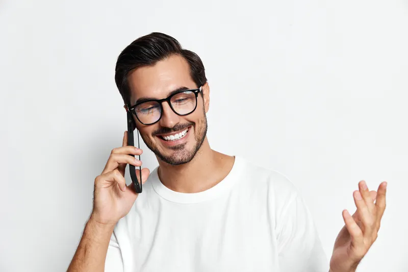 Smiling young man wearing glasses talking on a mobile phone with a cheerful expression, isolated on a plain white background. Modern lifestyle concept.