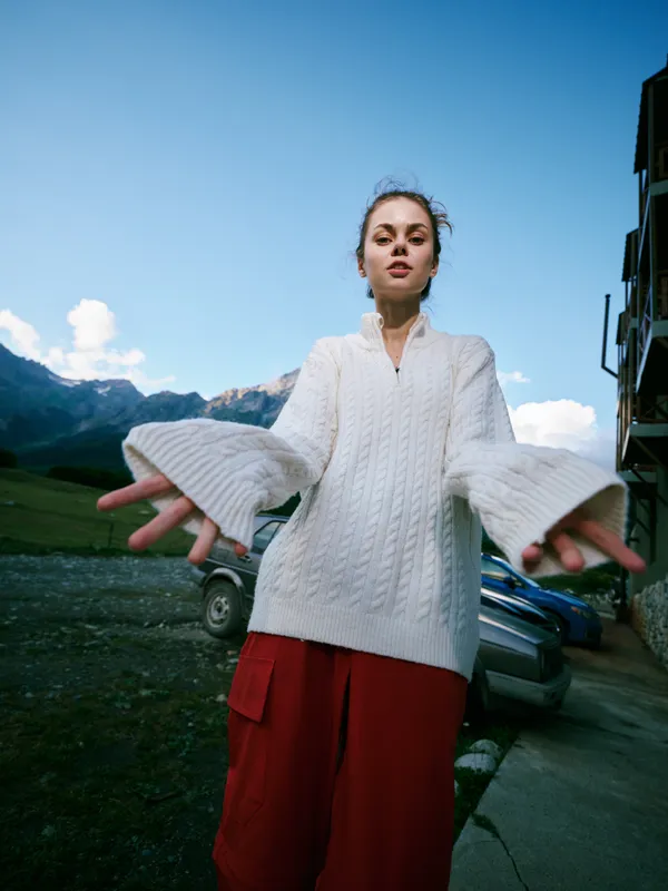 Outdoors fashion shot featuring a model in a white knitted sweater and red shorts, arms extended toward the camera with mountains and blue sky in the background for a bold, energetic look.